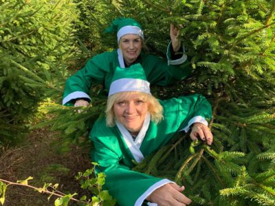 Two women, dressed as elves, hiding in some green bushes. They are smiling at the camera.