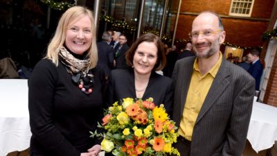 Three people, two women and one man, smilign at the camera. The woman in the middle is holding flowers.