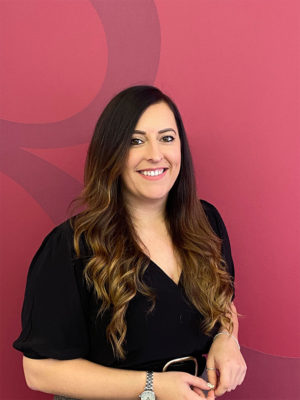 A woman with long dark hair, smiling at the camera. She is standing against a red wall.