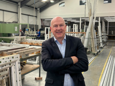 A smartly dressed man, folding his arms, smiling at the camera. He is standing in a large factory. 