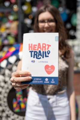 A woman with dark hair and glasses holding up a peice of paper t the camera. The paper reads 'Heart Trail'. 